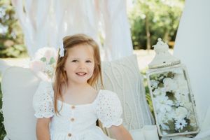 Child wearing white summer dress poses on cushions outdoors with decorative lantern.