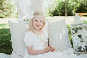 Young child in white eyelet dress sits on cushions in garden setting with decorative lantern.