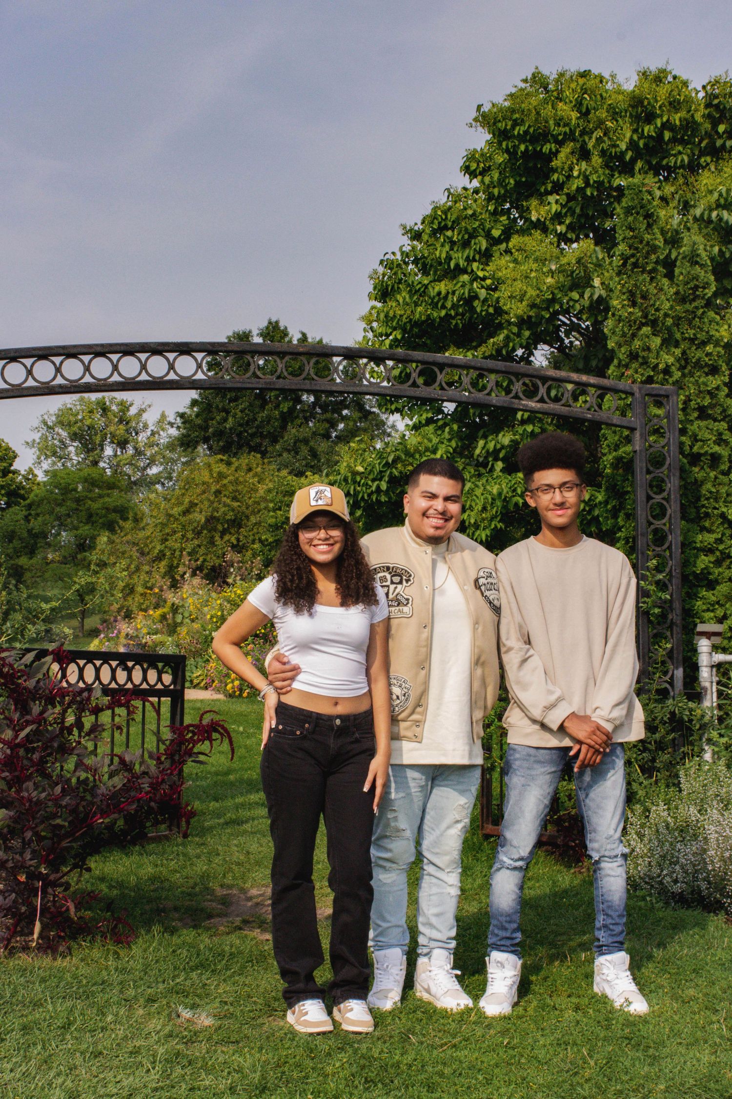 Three friends standing under garden archway in casual attire.