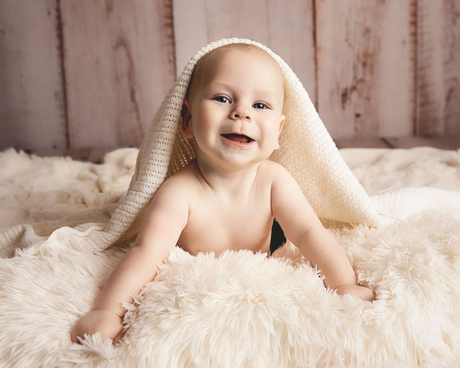 A smiling baby wrapped in a cream blanket lies on a soft white fur rug against a wooden backdrop.