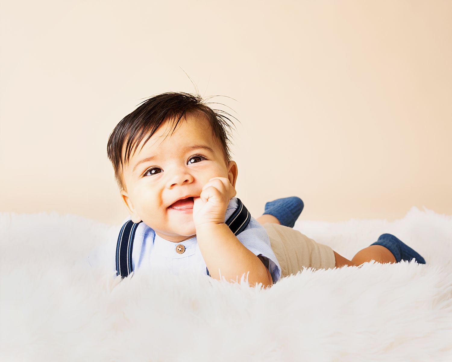 Baby in white onesie with blue trim lying on soft white carpet while smiling and touching their face.