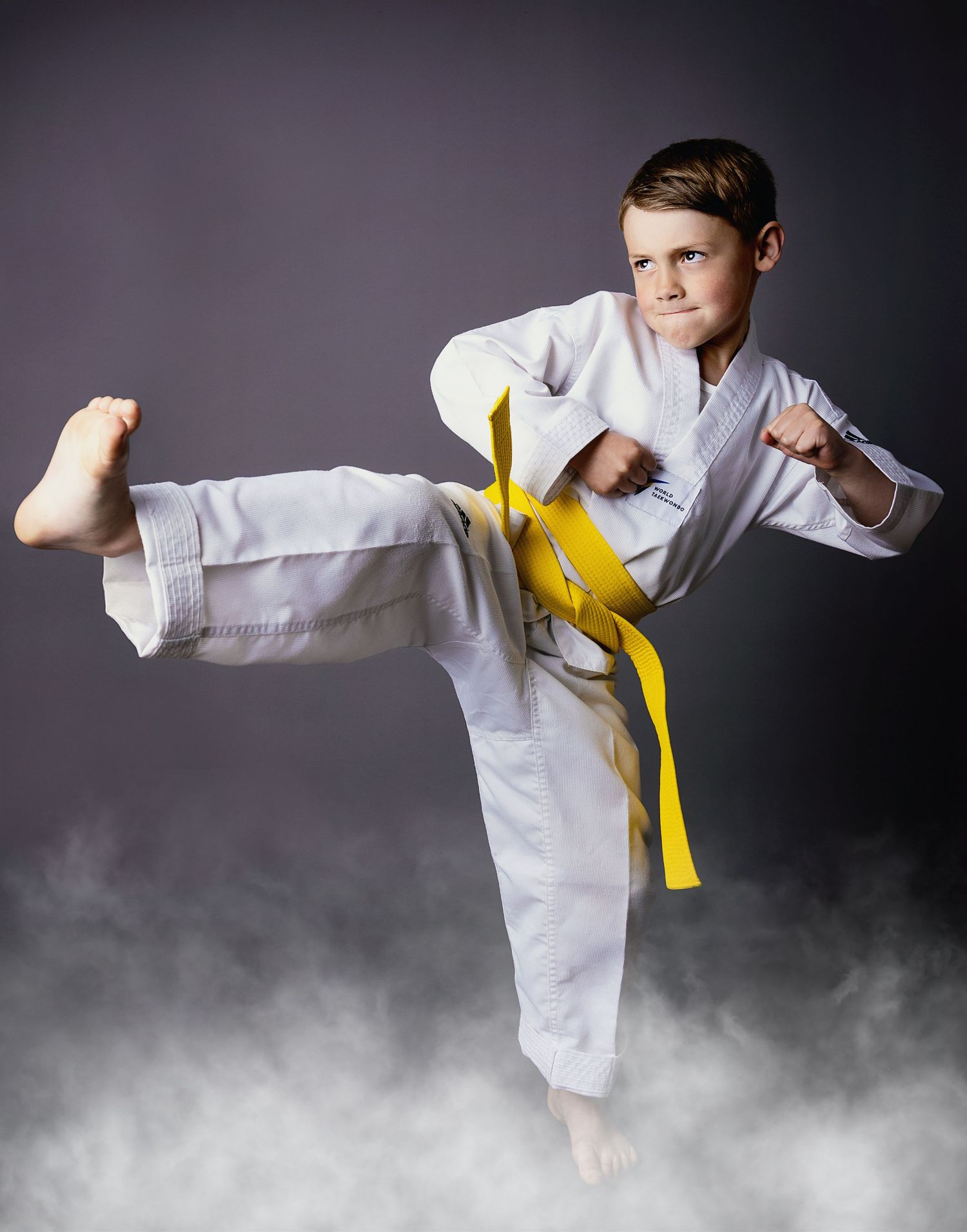 Young martial artist in white karate uniform with yellow belt executes a dynamic side kick in a smoky studio setting.