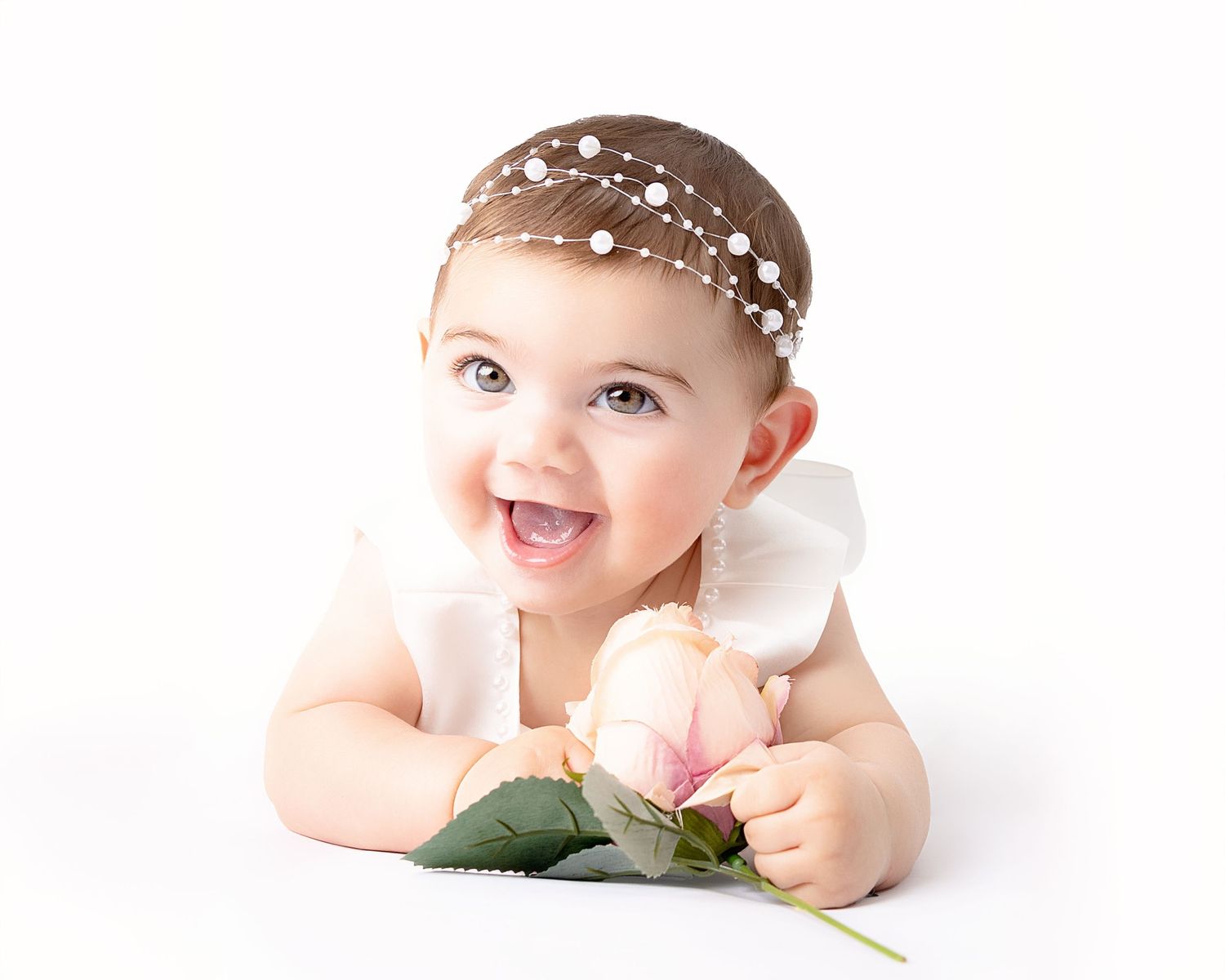 Baby wearing pearl headband lies on white backdrop holding pink rose flower with joyful expression.