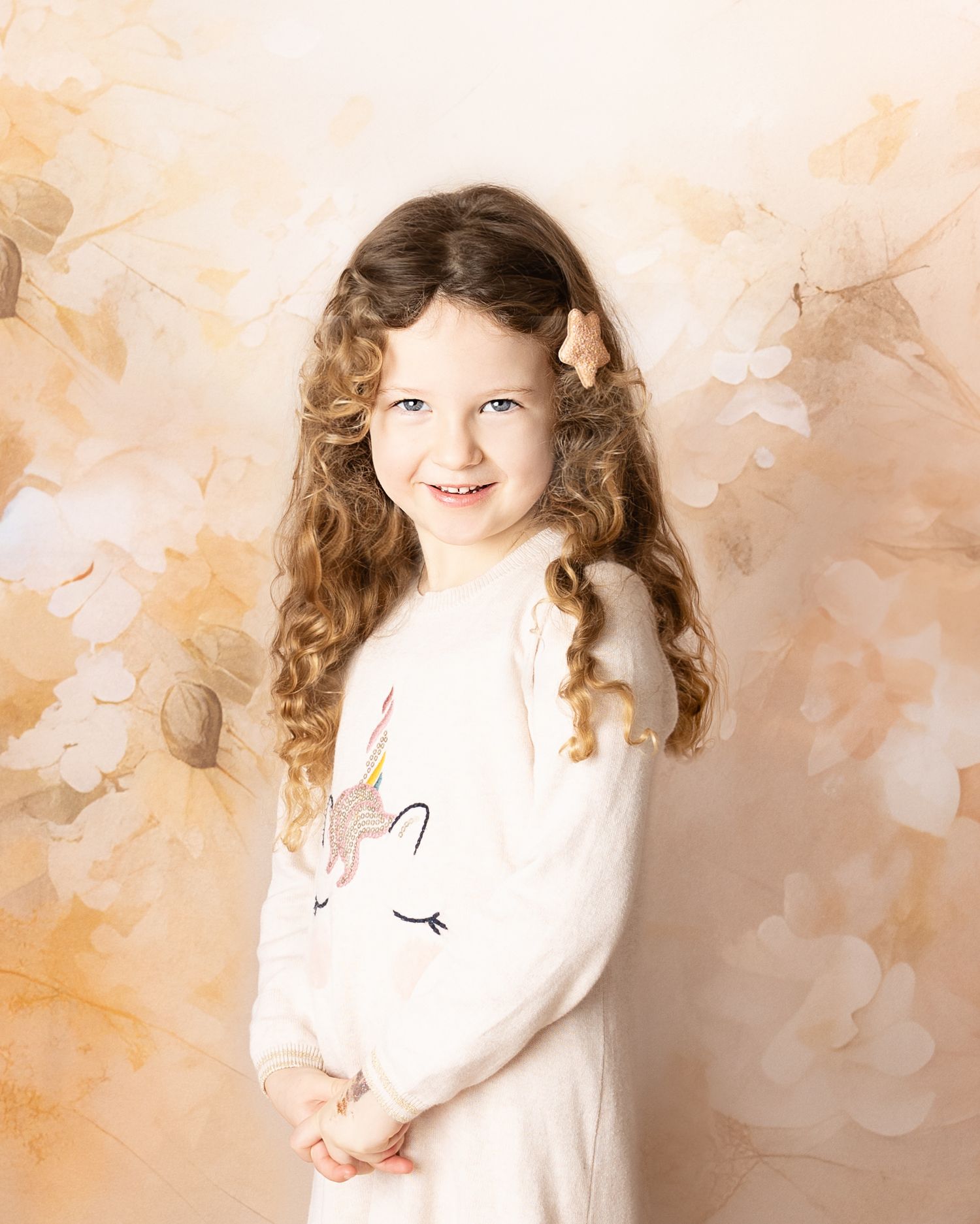 Portrait photograph with a floral background featuring long curly hair and white clothing in warm sepia tones.