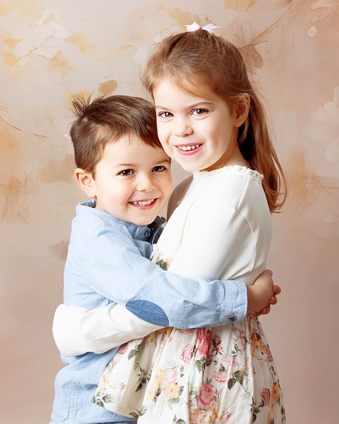 Two young children in coordinated blue and white outfits embrace warmly against a neutral studio backdrop.