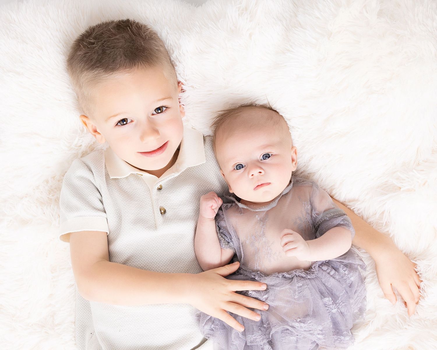 Two young children wearing light colored clothing lay together on a white background, looking up at the camera.