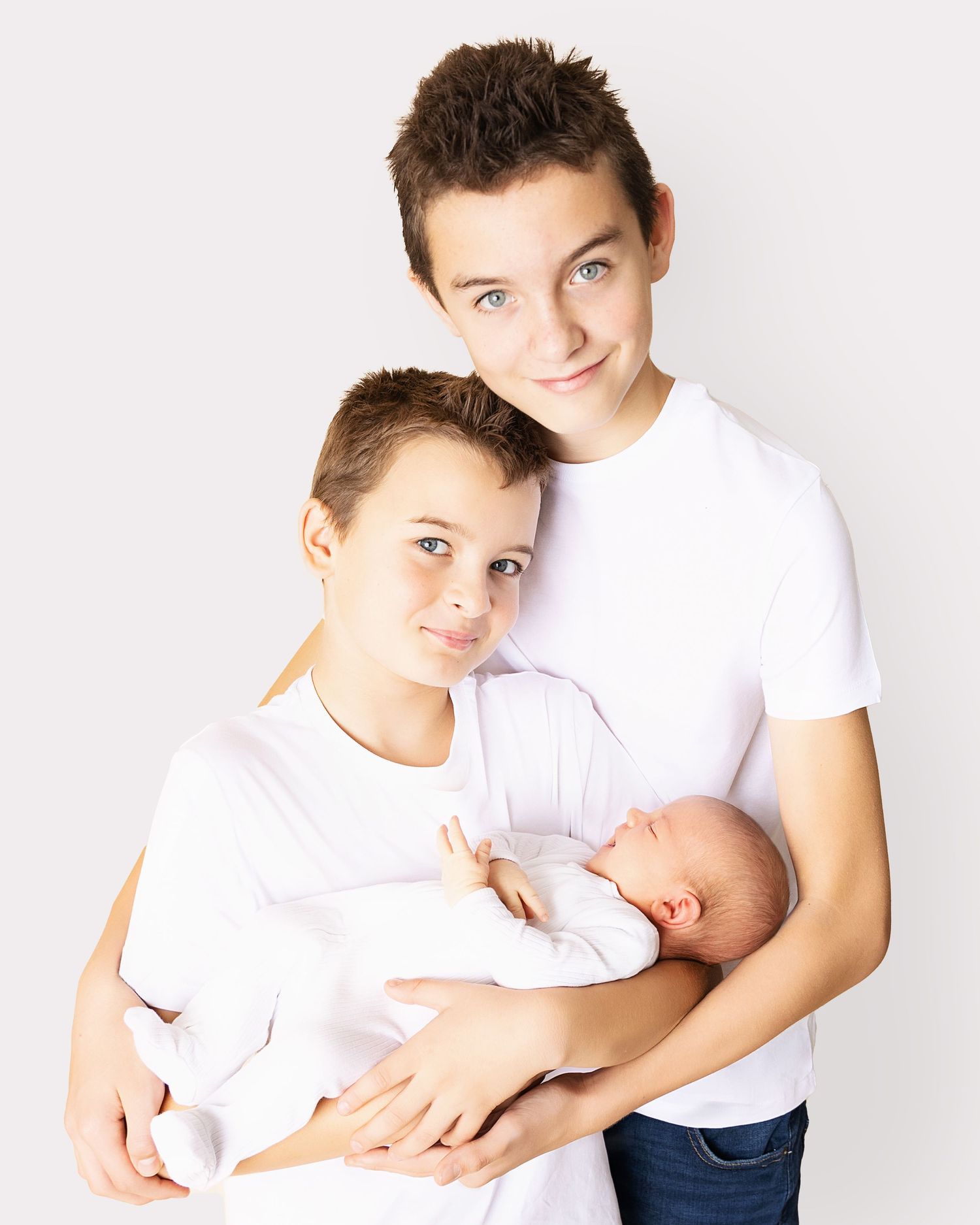 Three siblings dressed in white clothing posed together, with an infant being cradled by the older pair on a light background.
