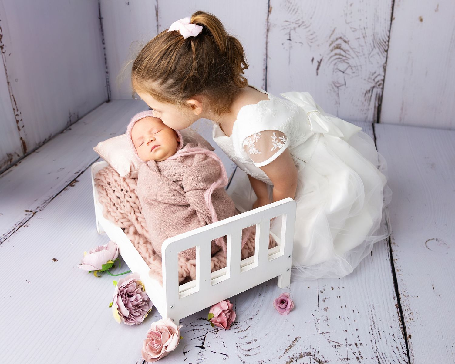 A figure in a white dress leans over a small white bed containing a doll with pink roses scattered around on a white floor.