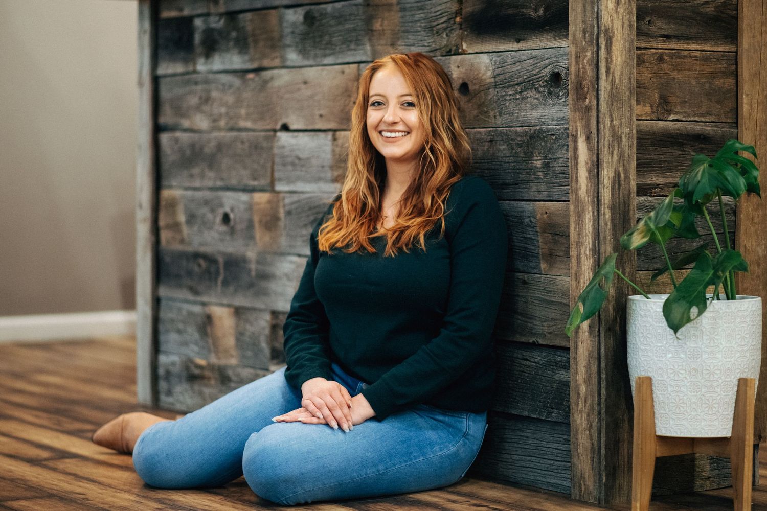 Woman in dark sweater and jeans sits casually on wooden bench against rustic wall with indoor plant nearby.