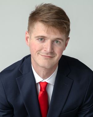 Professional headshot portrait of someone in a navy suit and bright red tie against a light background.