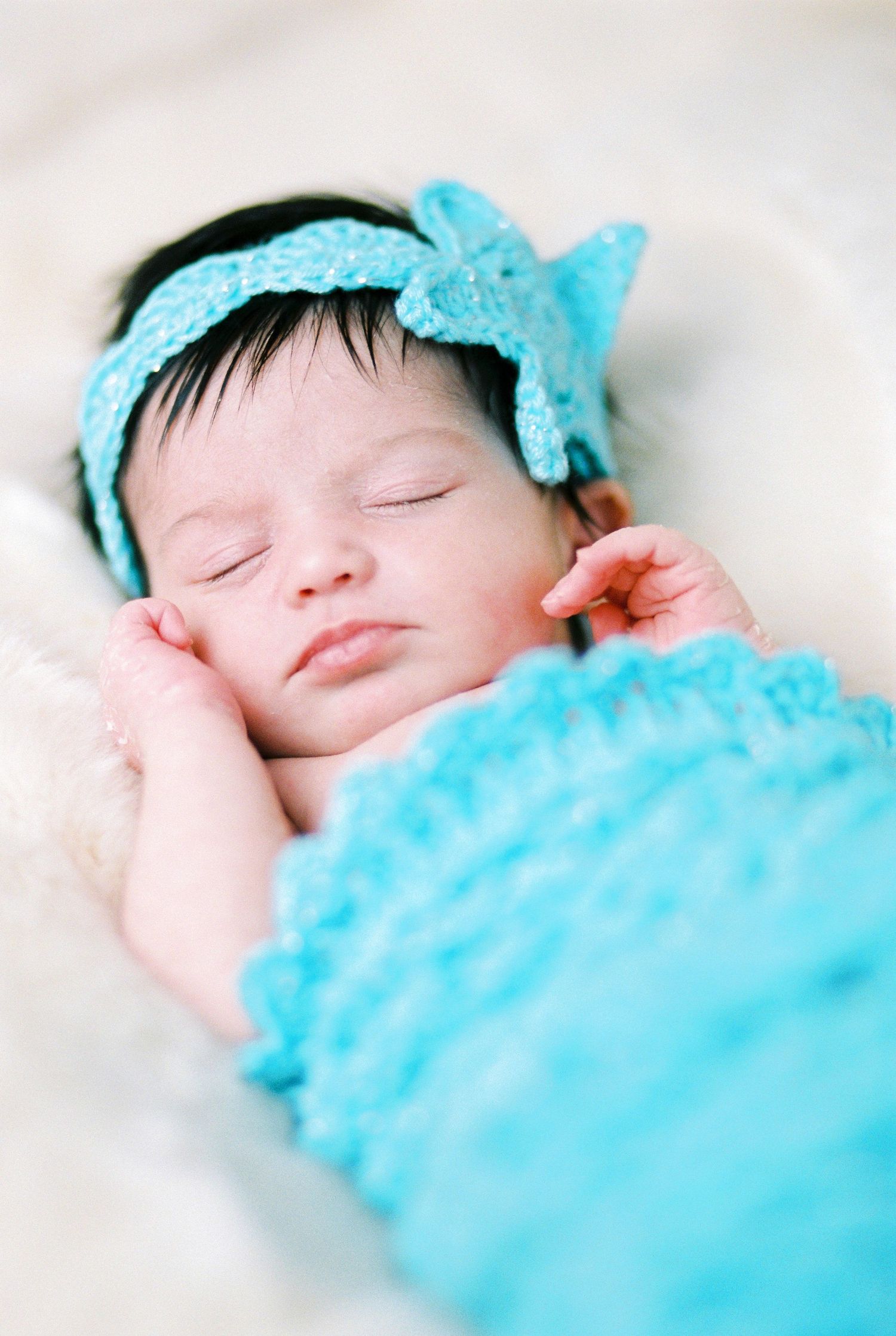 Sleeping newborn wearing turquoise bow headband and wrapped in matching turquoise fabric.
