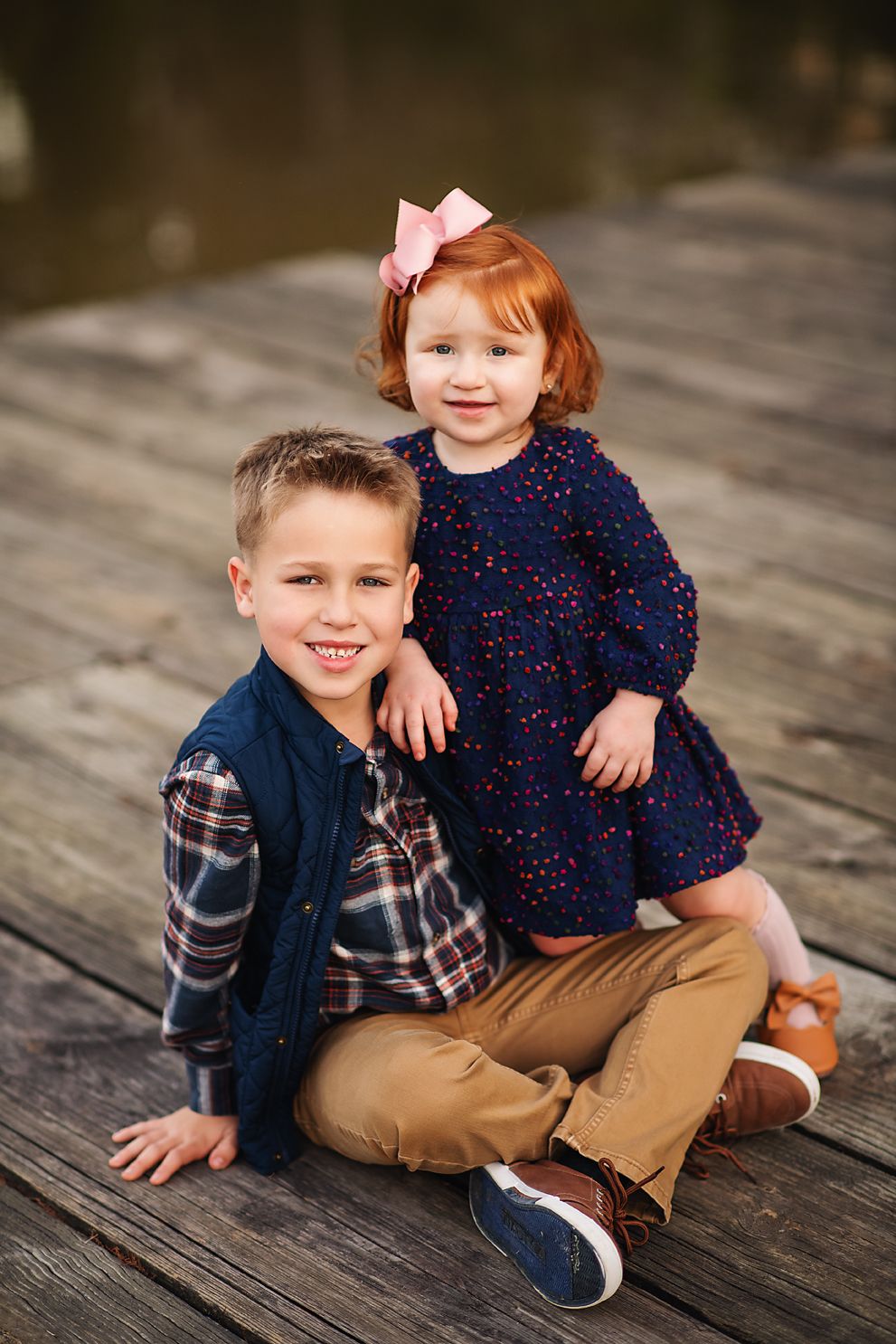 Young boy and toddler girl with red hair and pink bow sitting together on a wooden dock smiling.