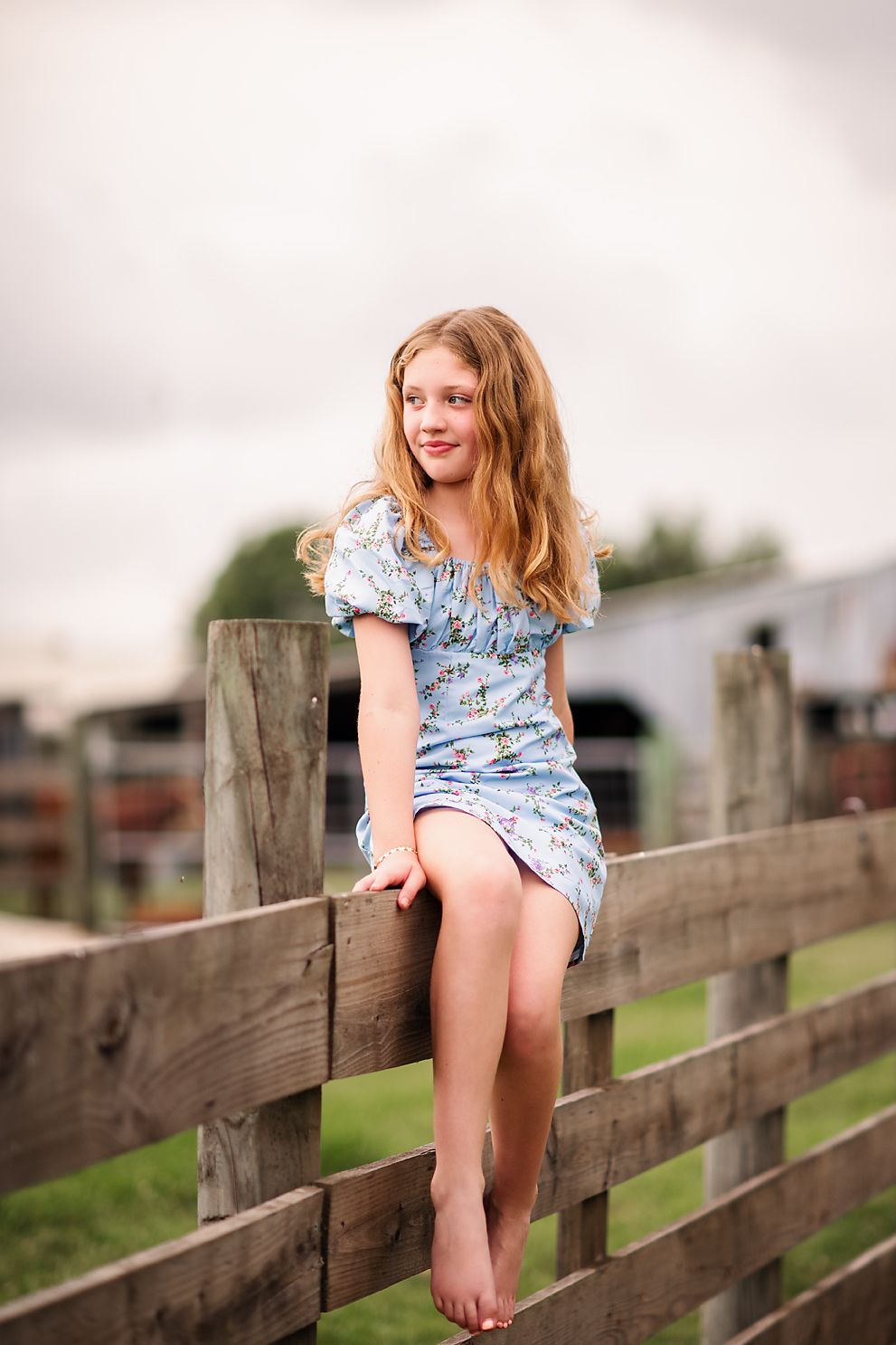 Young girl in floral blue dress sitting barefoot on wooden farm fence with green pasture background.