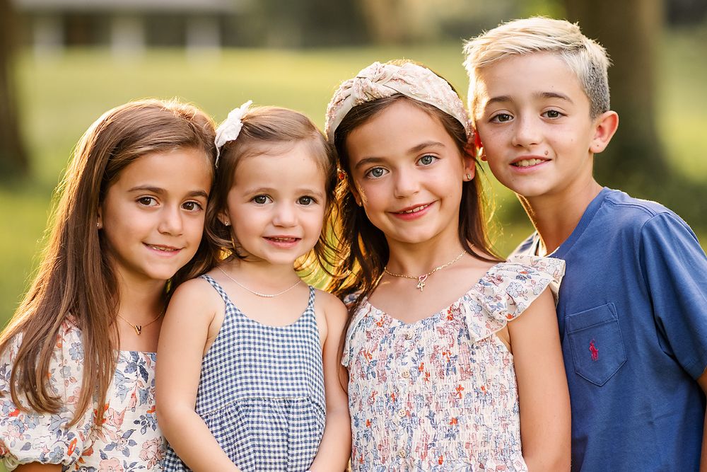 Four smiling children posing outdoors in a sunny park, wearing summer outfits, capturing a joyful family moment.