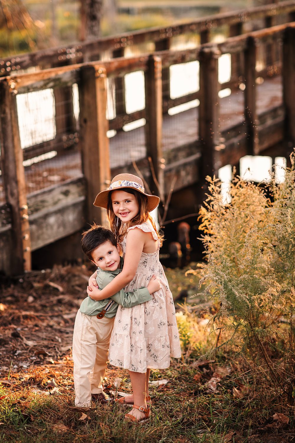 Smiling girl in floral dress and hat hugging younger brother near rustic wooden bridge at sunset.