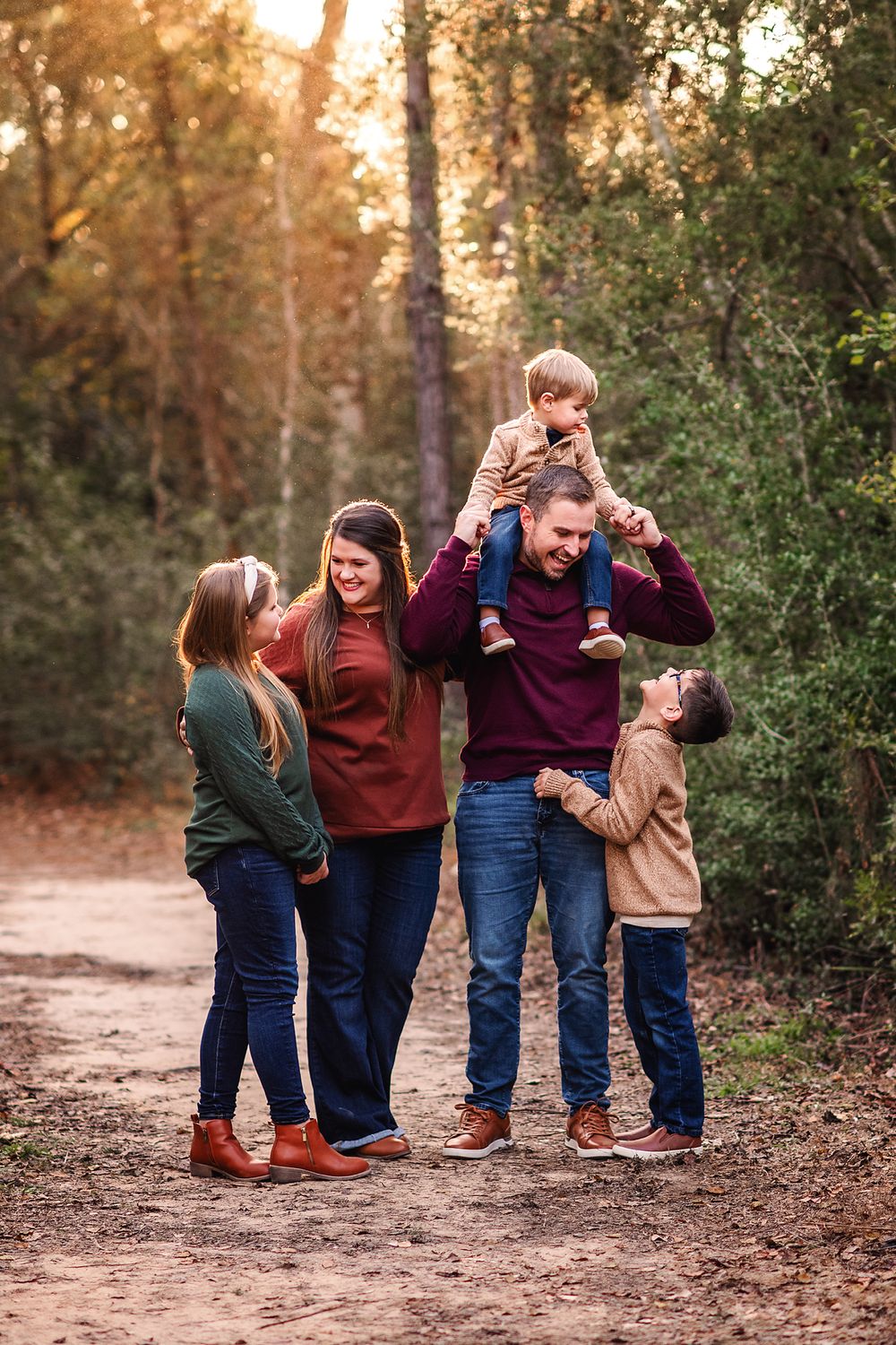 A joyful family of five plays on a forest path, dad lifting toddler on shoulders at golden hour.