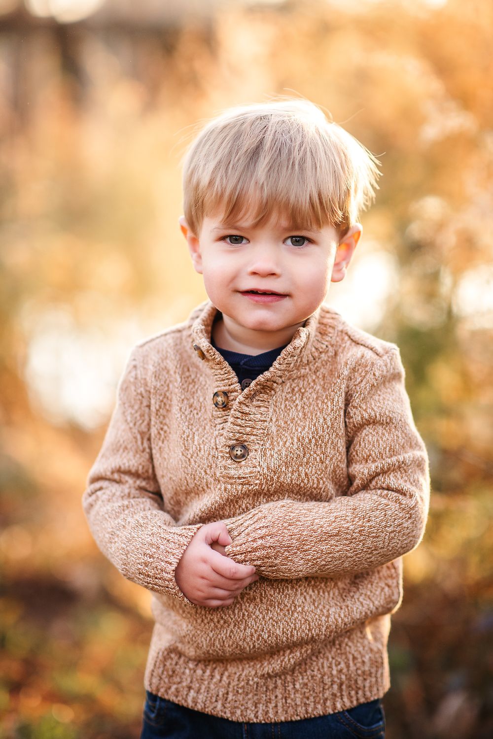 An adorable blonde toddler boy in a beige knit sweater smiles outdoors in autumn light.