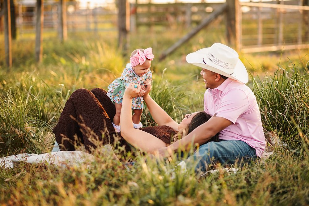 Family sitting in grass with parents playing with baby girl wearing pink bow outdoors at golden hour.