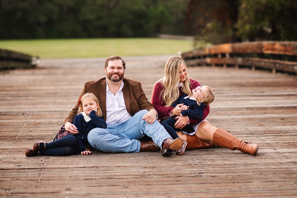 Happy family sitting on a wooden dock outdoors in fall, parents smiling with two young children in coordinated outfits.