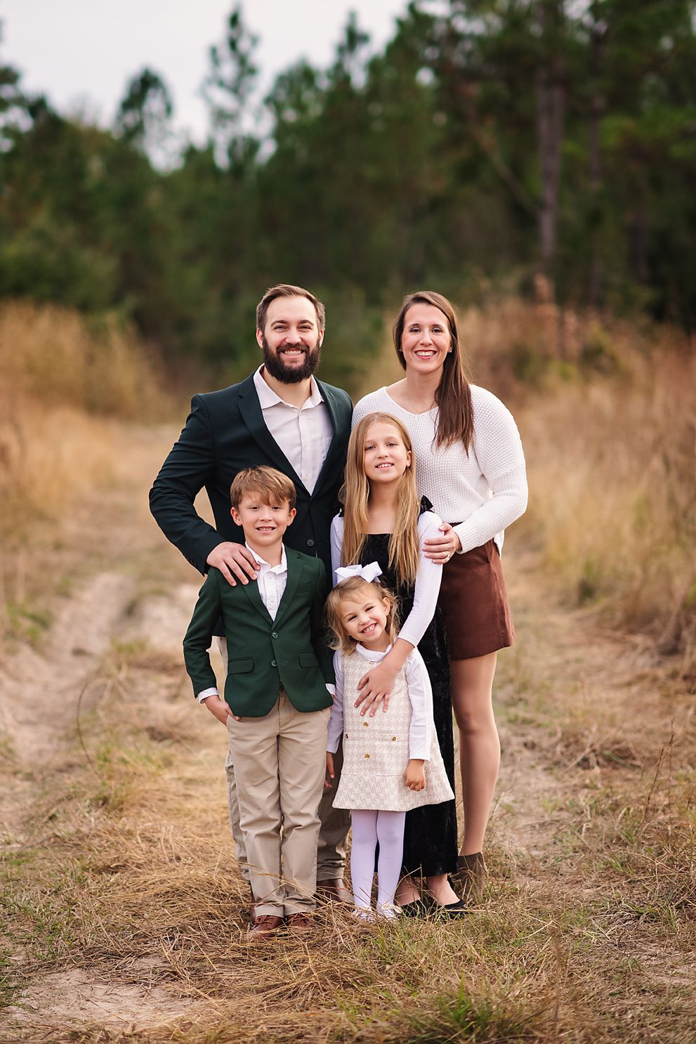 Happy family of five posing on a dirt path surrounded by tall grass and pine trees.