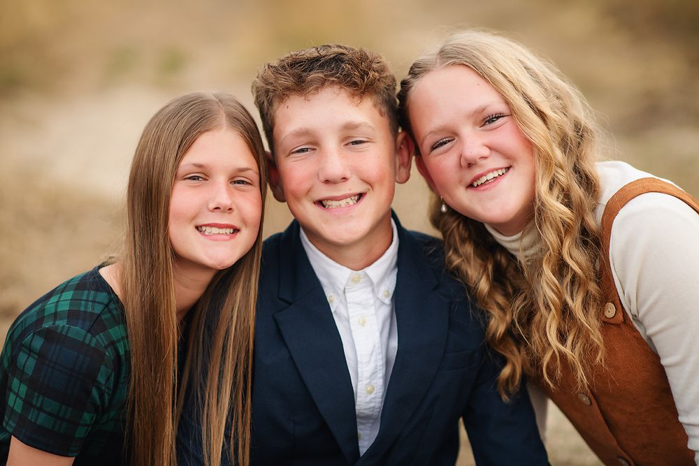 Three smiling siblings posing together outdoors, two girls and a boy in semi-formal attire.