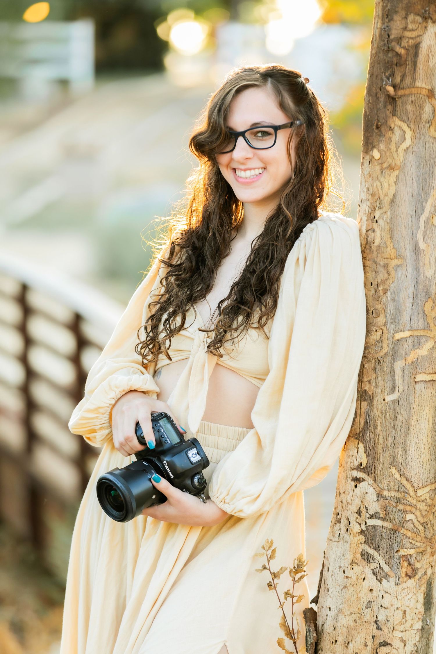 Photo of Victorville Ca Family photographer Ashley Ann Imagery leaned against a tree while holding a camera in her hands smiling