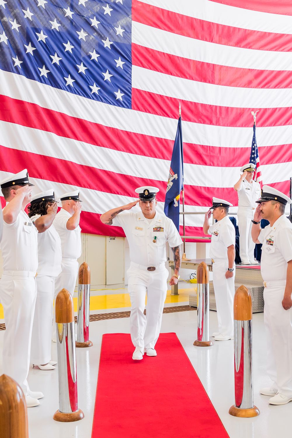 Naval service members stand at attention during a military ceremony with a large American flag backdrop.