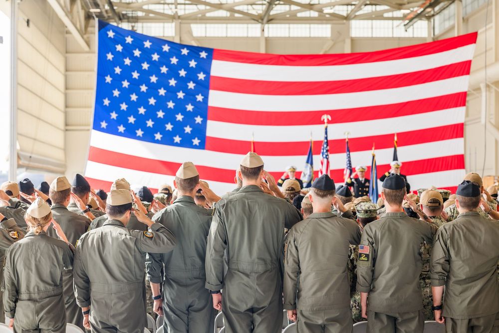 Military personnel stand in formation at attention while American flags hang prominently in the background.