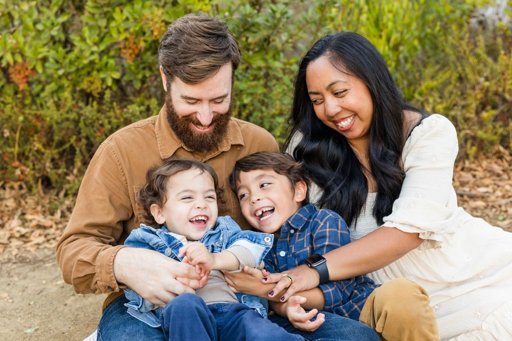 A joyful candid family portrait showing parents and two young children hugging and laughing together outdoors.