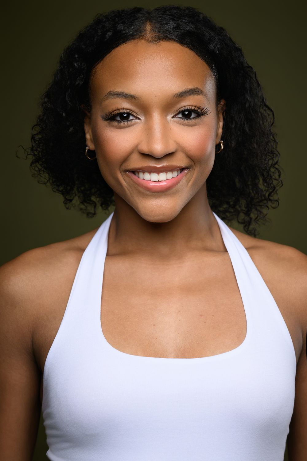 Smiling young Black woman with curly hair wearing a white halter top against a dark green background.