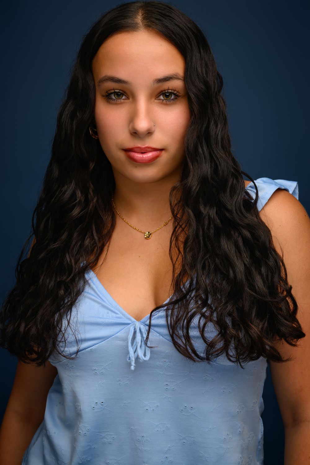 Young woman with long wavy dark hair wearing a light blue top and gold necklace poses against a dark blue background.