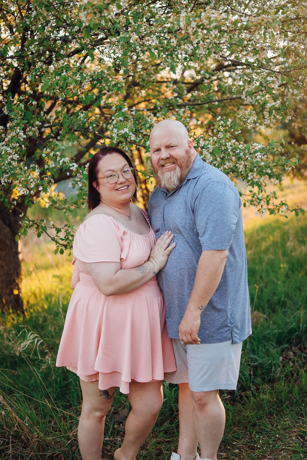 A smiling couple poses under a blooming tree at golden hour, woman in pink dress, man in gray shirt.