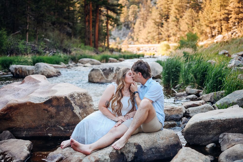 Couple sharing a romantic kiss while sitting on rocks beside a mountain stream surrounded by lush greenery.