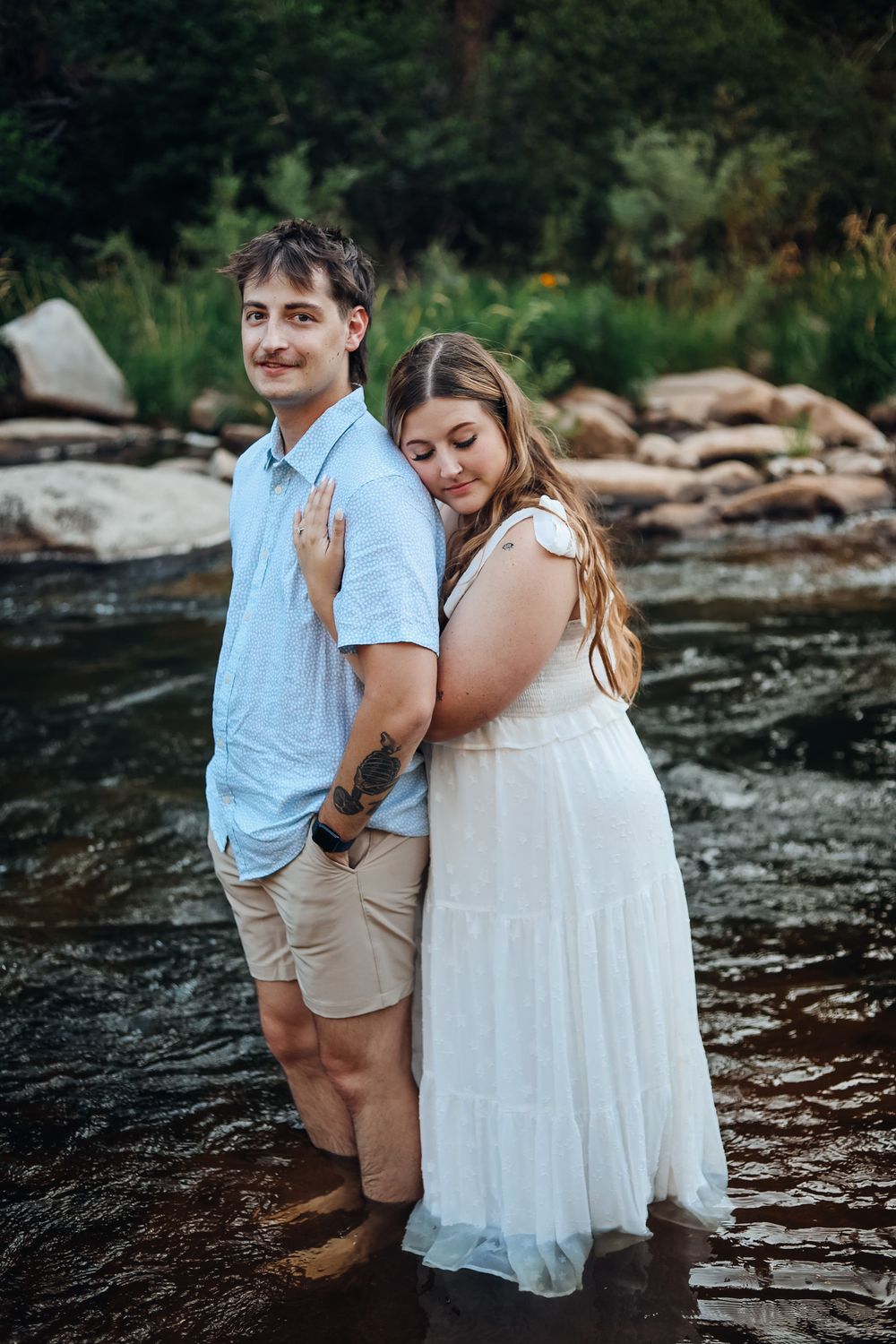 Couple standing in a shallow river, woman in white dress embracing man in blue shirt and khaki shorts, with rocks and greenery behind.
