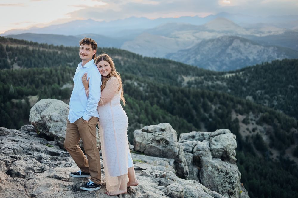 Couple posing on rocky mountain summit with scenic forest and mountain range backdrop at sunset.