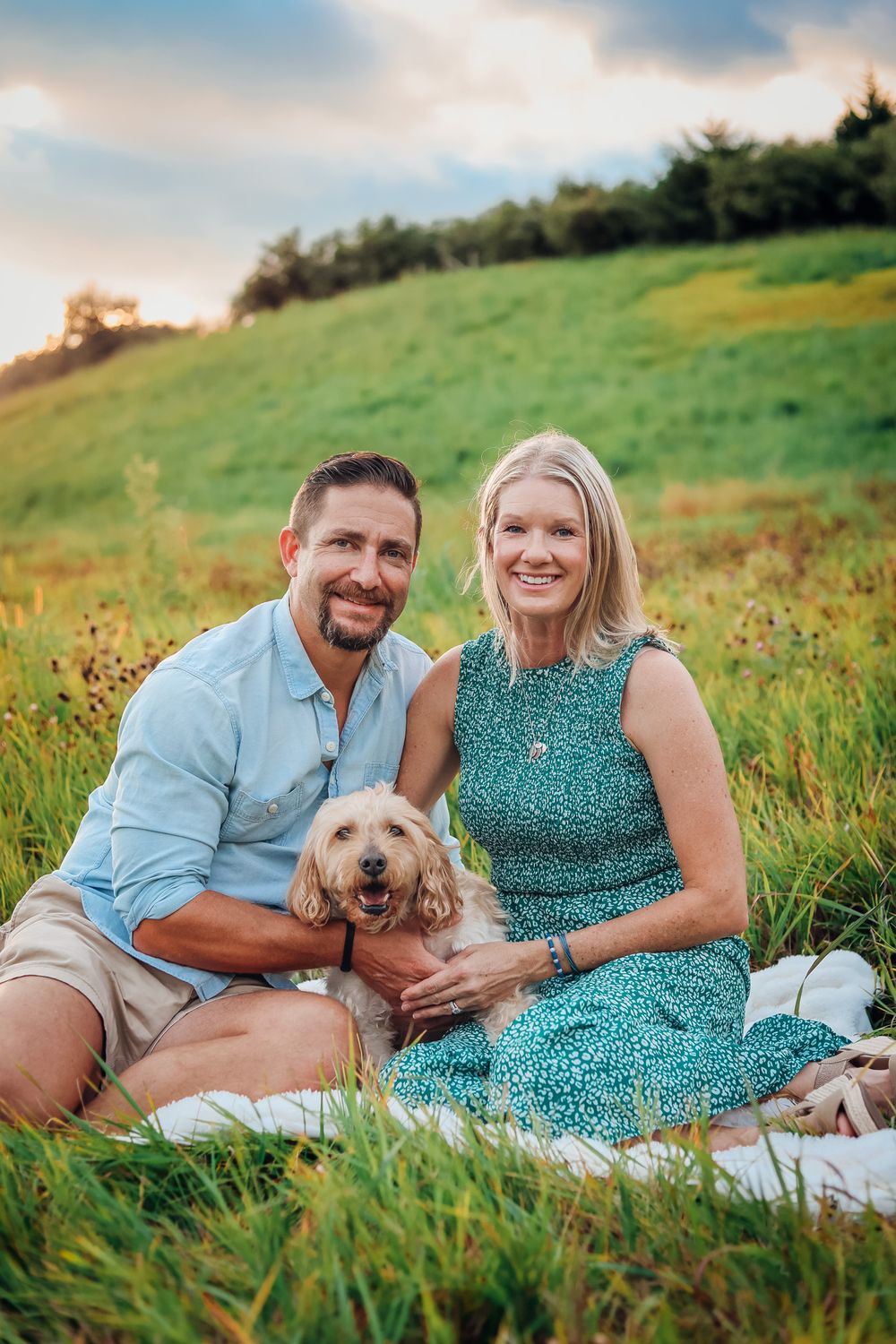 Smiling couple sitting on grass with golden doodle dog during sunset picnic in green meadow.