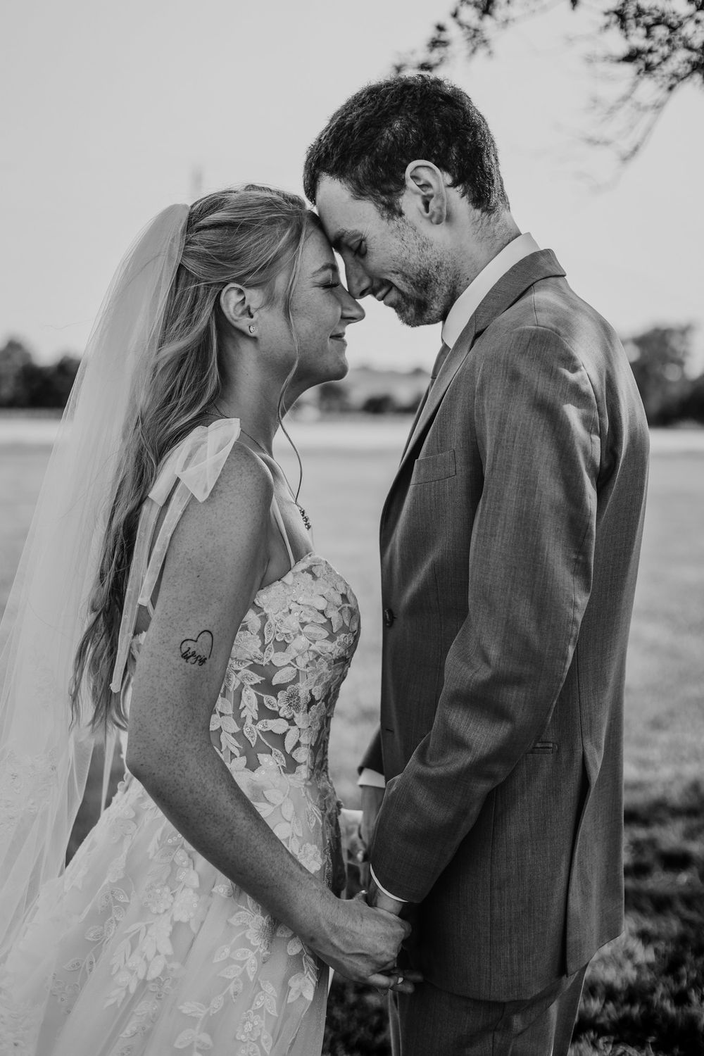 Black and white wedding photo of bride and groom holding hands, foreheads touching, smiling outdoors.