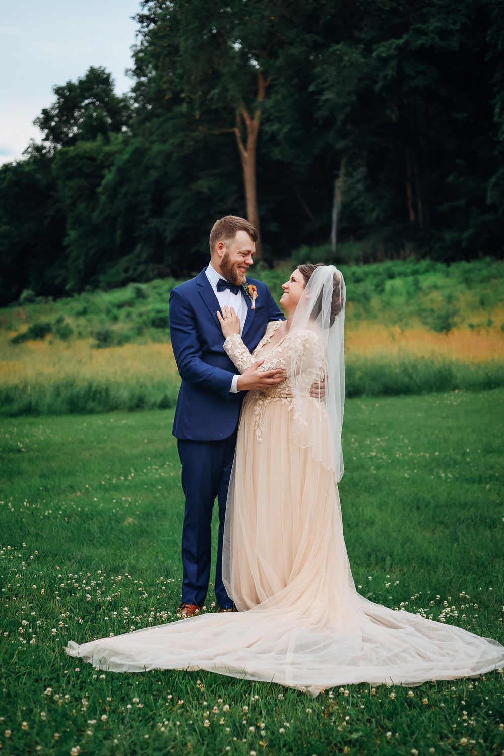 A bride in a champagne gown and veil smiles with a bearded groom in a blue suit on a green lawn.