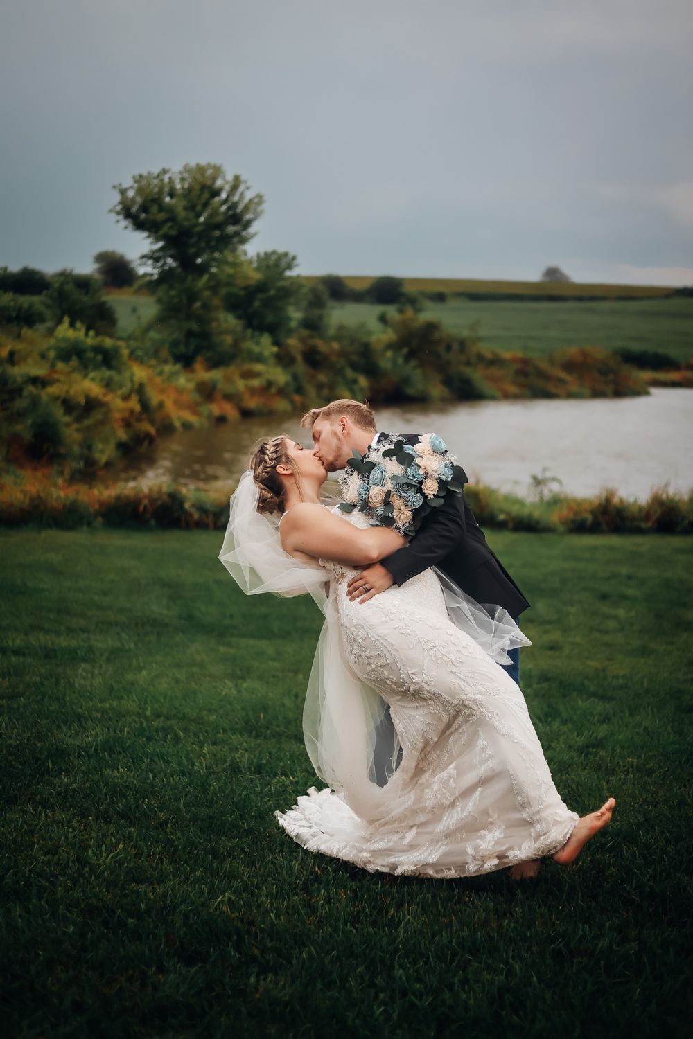 Groom dips bride for a romantic kiss on lush green lawn with scenic lake and trees in background at outdoor wedding.