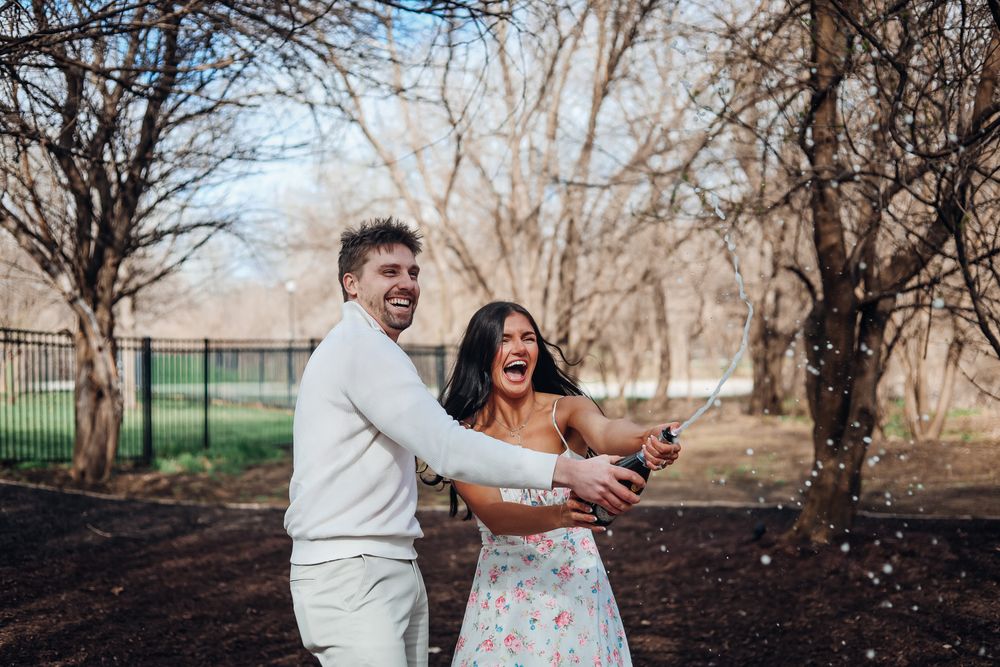 Couple laughing and popping champagne bottle outdoors among bare trees in a joyful engagement celebration photo.