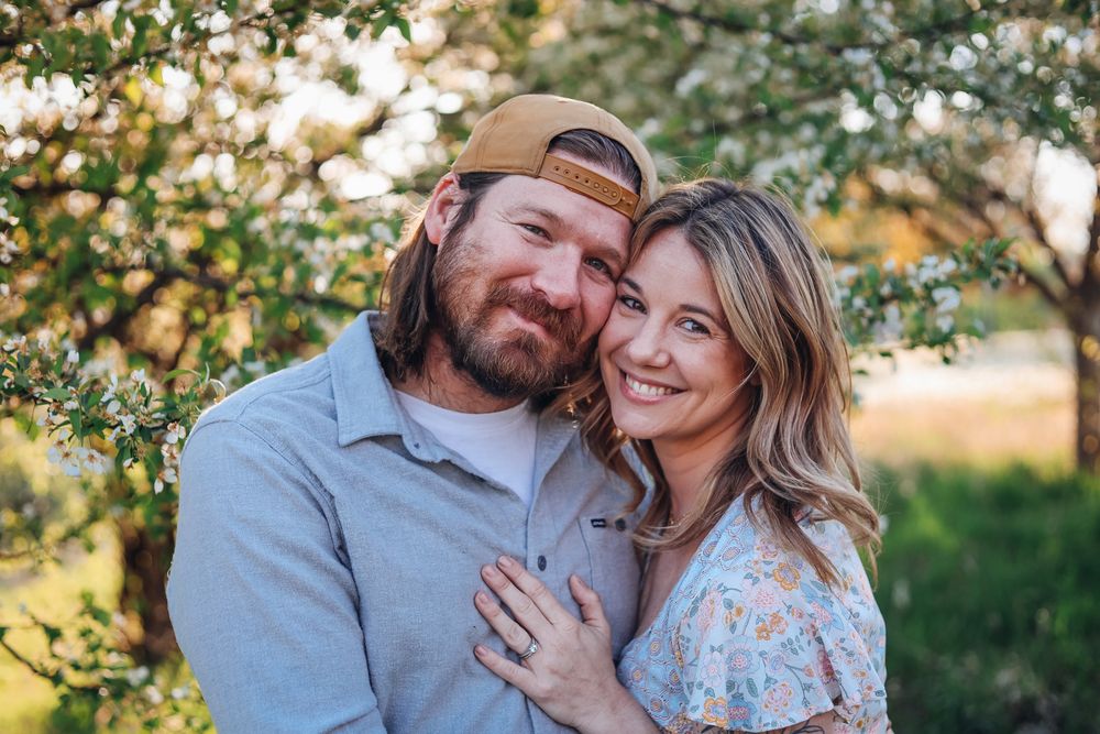 A smiling couple embraces outdoors under blooming white trees in warm golden light, the man wearing a backward cap.
