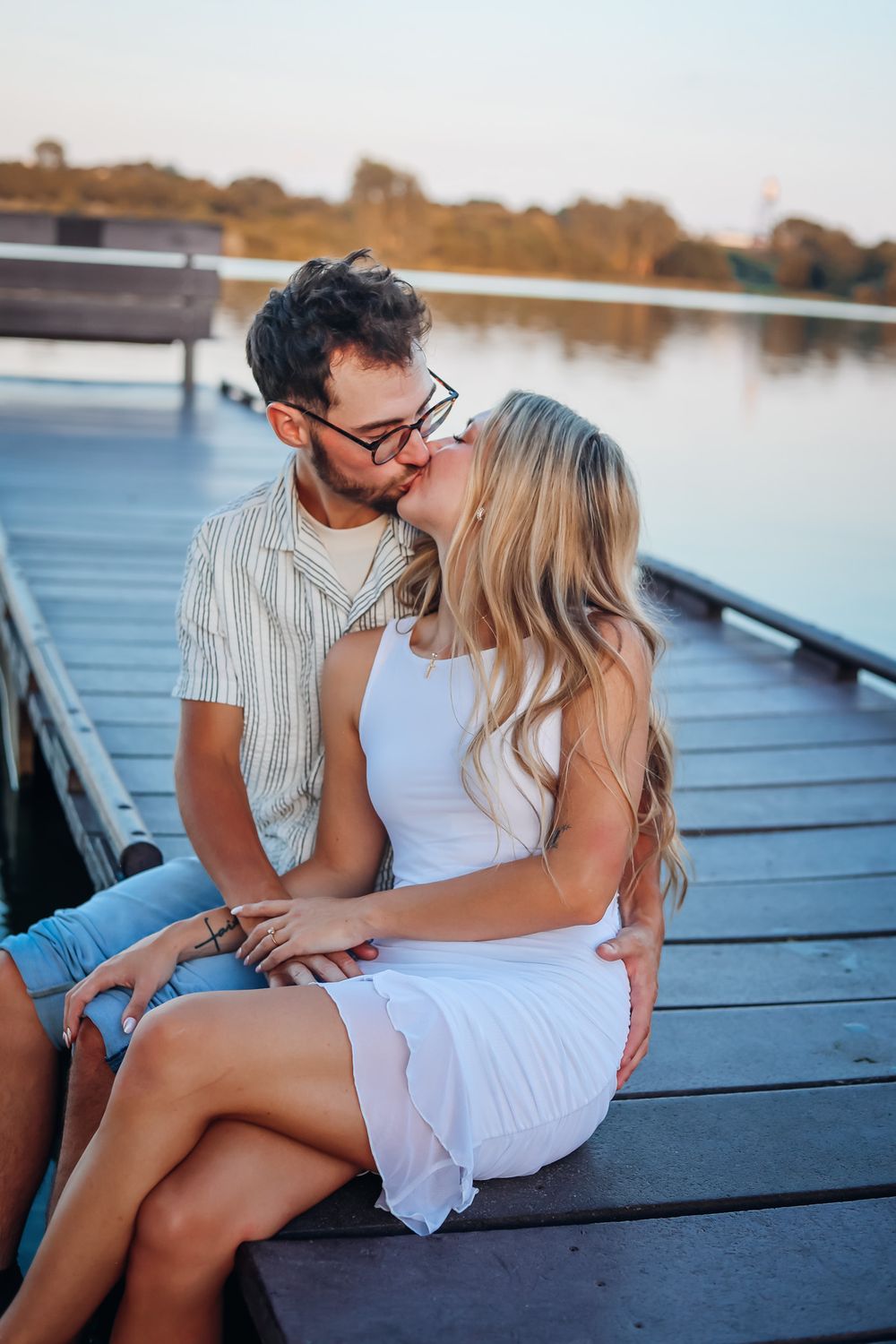 Couple sharing a romantic kiss on a wooden dock by a calm lake at sunset, woman in white dress.