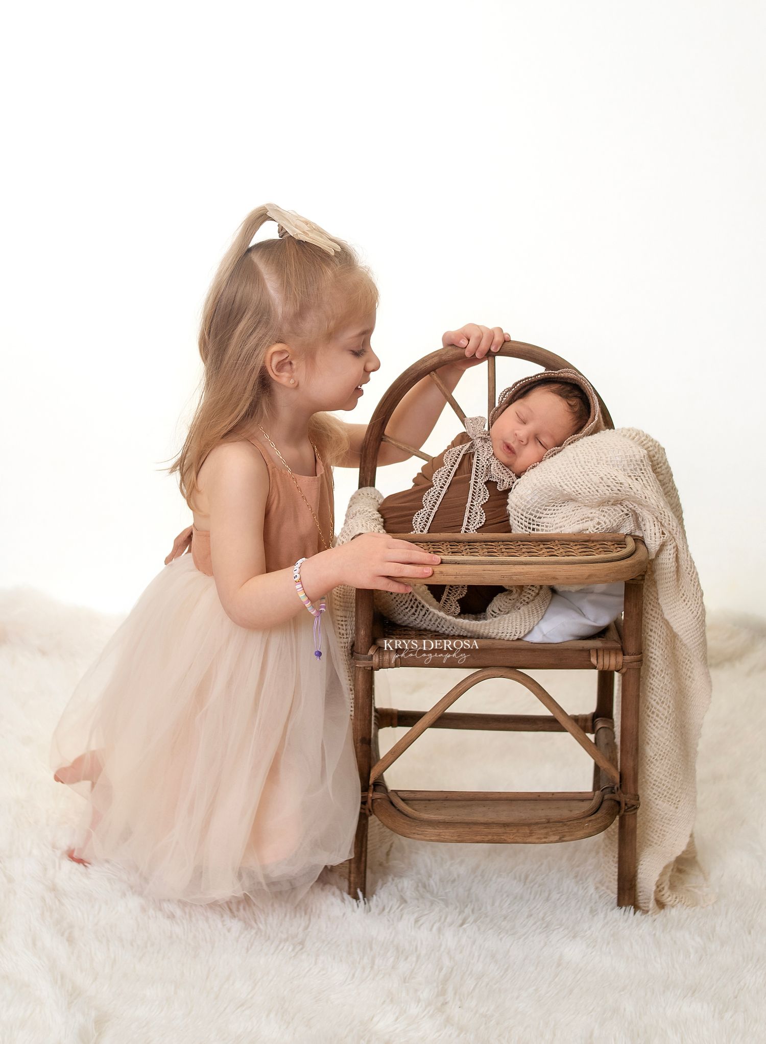 Young girl in a peach tulle dress kneels beside newborn sibling in an antique woven chair with rustic wheel design in studio setting.