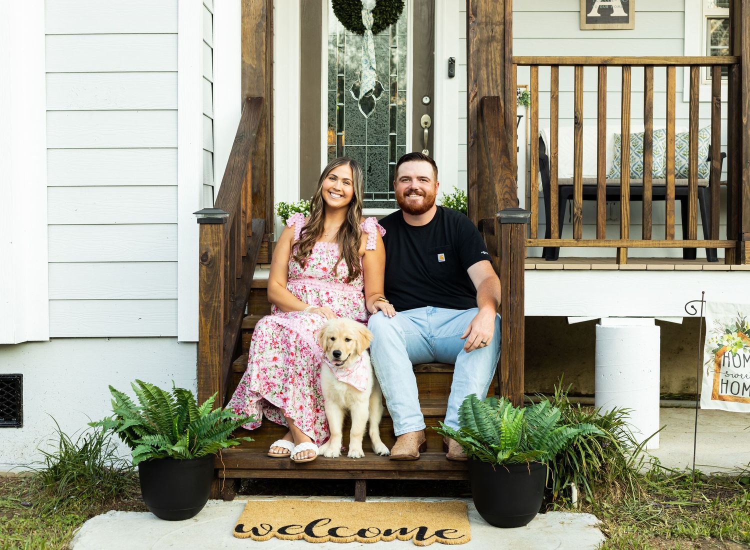 couple sitting on a front porch with a dog and welcome mat.