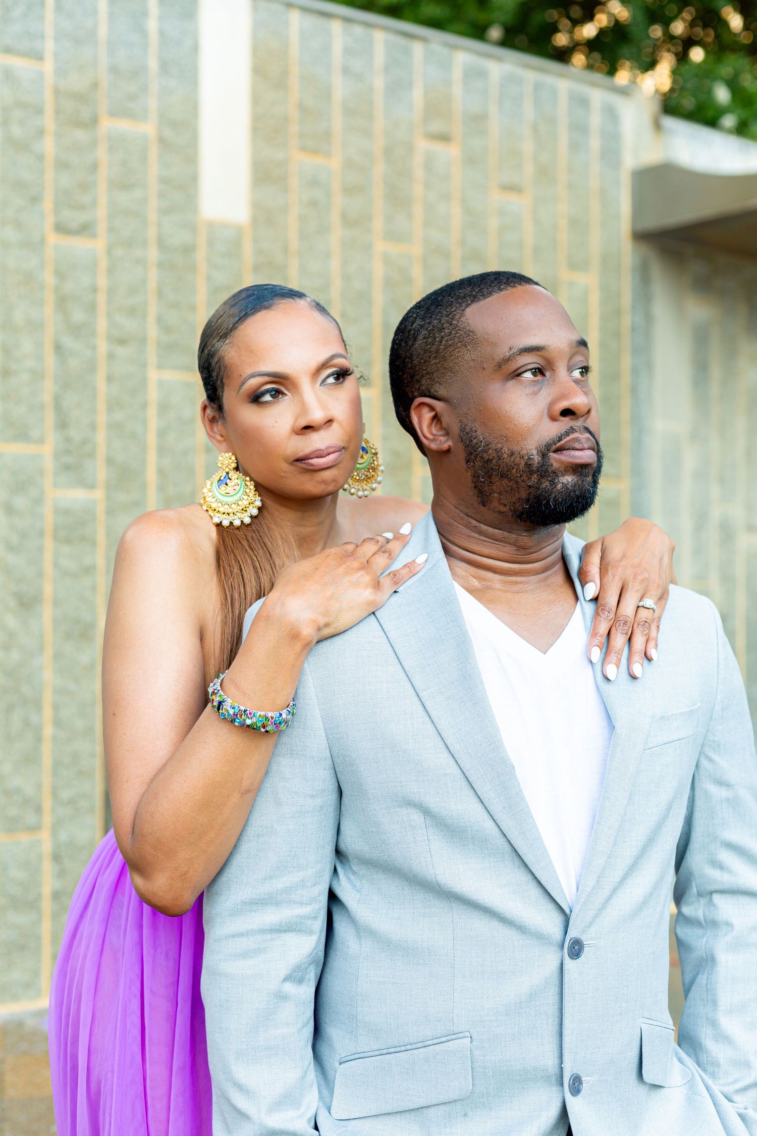 Couple poses outdoors in formal attire with woman in purple dress and man in light gray blazer.