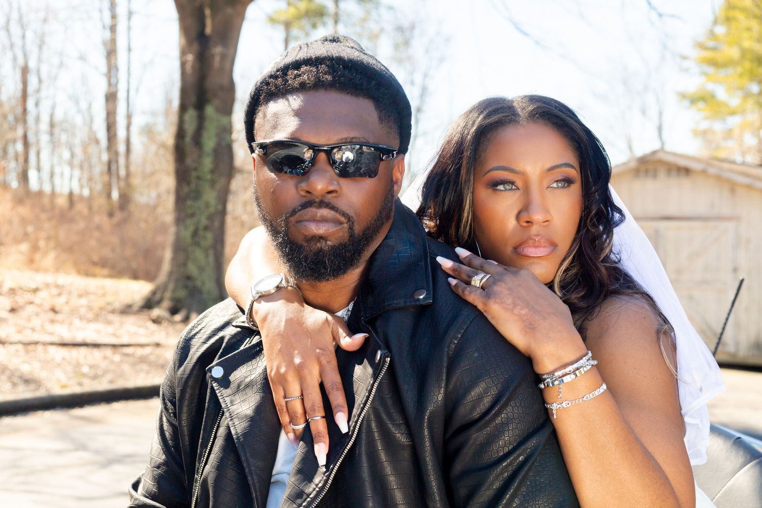 A stylish couple poses together outdoors, wearing black leather and sunglasses in warm sunlight.