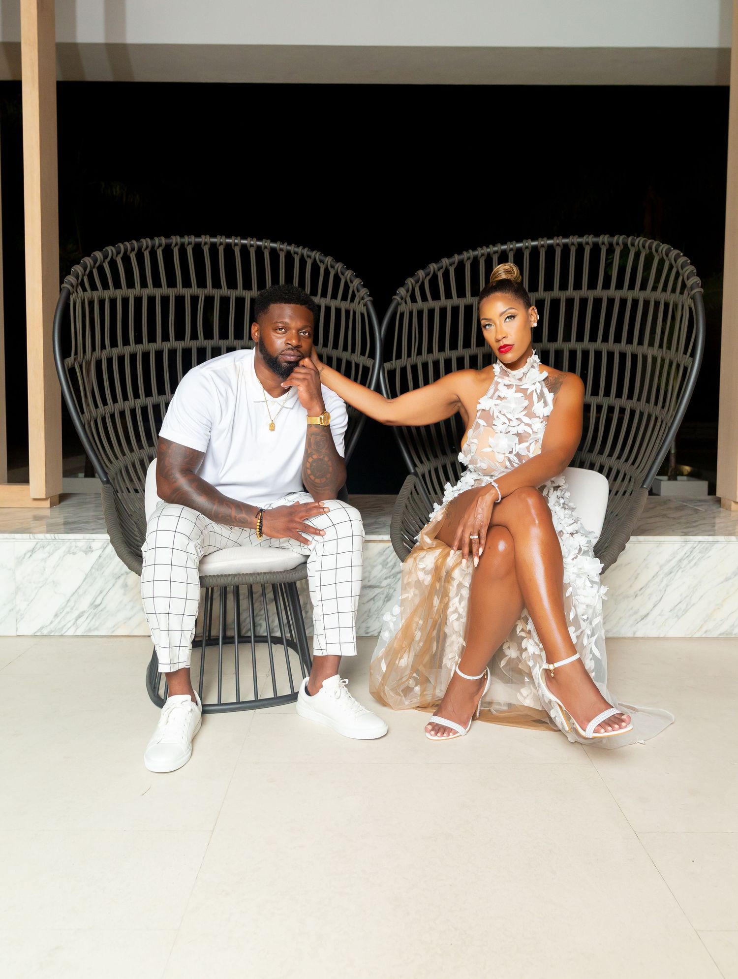 Two people relax together on wicker peacock chairs on a porch, dressed in stylish white and cream outfits.