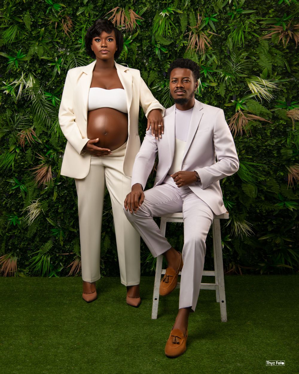 A couple wearing matching cream-colored suits poses for a maternity photo against a lush tropical plant backdrop.