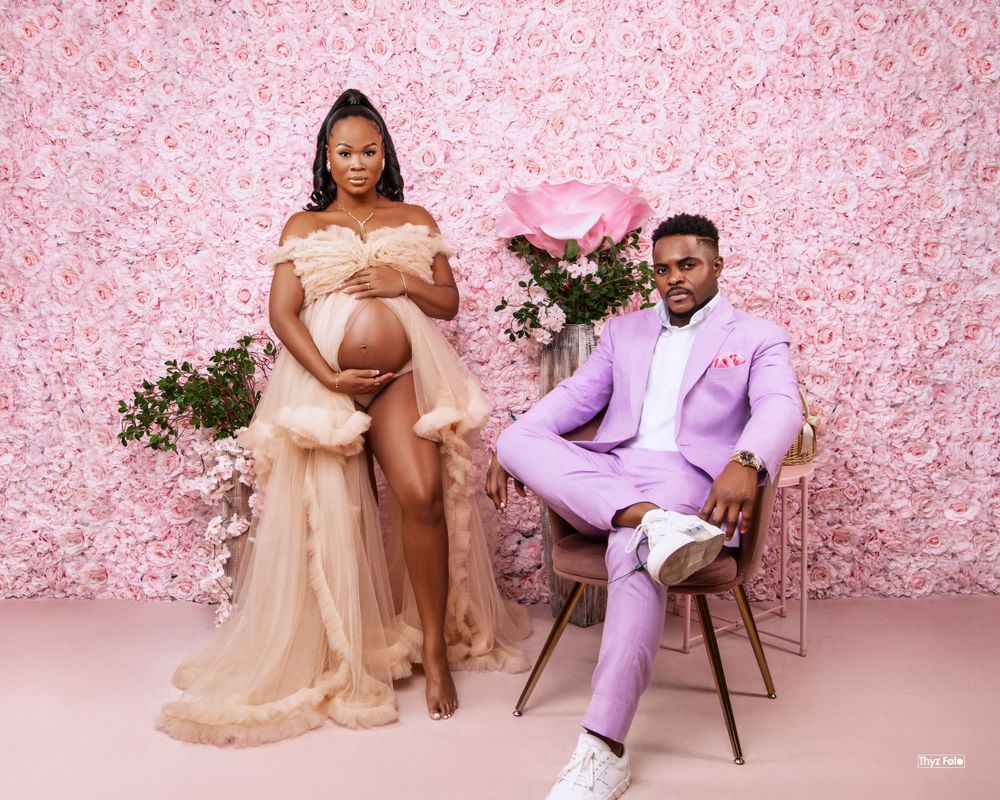 Maternity photoshoot against pink floral backdrop features couple in formal attire with lavender suit and champagne gown.