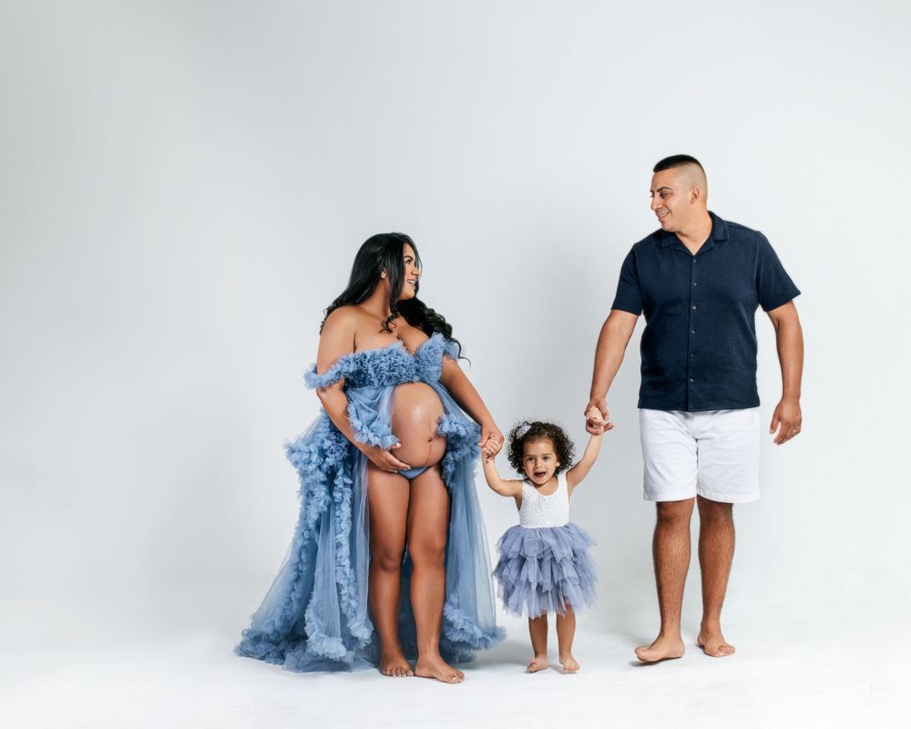 Beautiful family maternity portrait featuring a pregnant mother in a flowing blue gown, accompanied by her partner and child against a white
